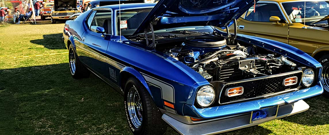 A bright blue 1960s muscle car is parked at a car show with the hood open.