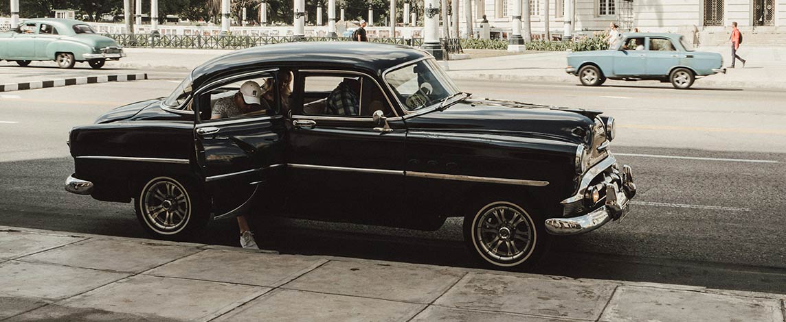 A passenger enters an elegant black 1950s car parked on the curb.