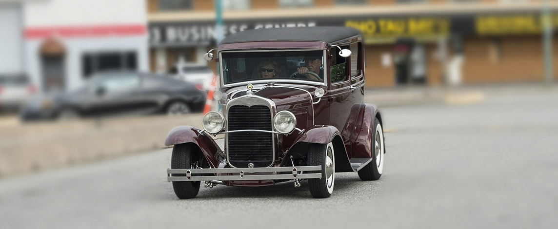 A 20th century maroon car cruises down the street.